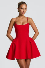 A woman modeling a bright red sleeveless mini dress with thin shoulder straps, a fitted bodice, and a flared skirt against a light gray studio background.