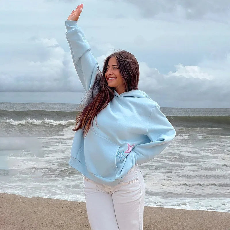 Woman in light blue hoodie and white pants standing on a beach with ocean waves in the background