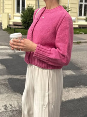 Person wearing a pink knitted cardigan and white pants, holding a coffee cup outdoors.