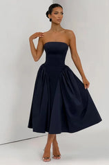 A woman modeling a strapless midi dress in dark navy blue. The dress features a structured, fitted bodice and a voluminous, pleated skirt.