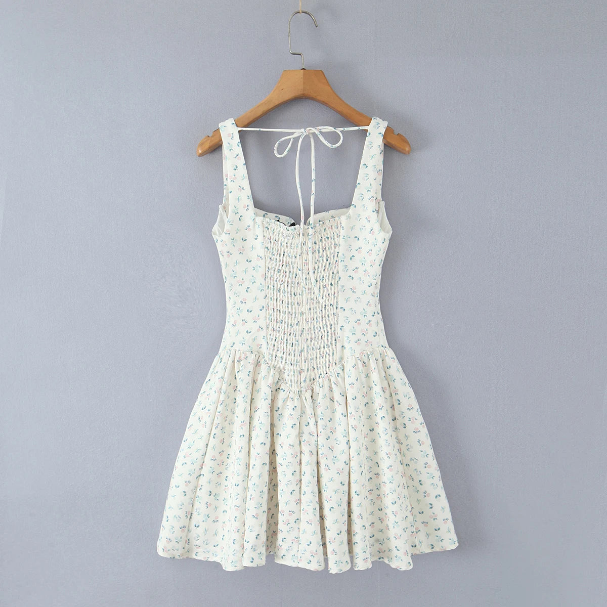 A sleeveless white smocked dress with a tiny blue and green floral print, hanging on a wooden hanger against a solid gray wall.