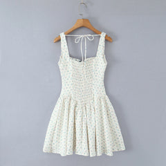 A sleeveless white smocked dress with a tiny blue and green floral print, hanging on a wooden hanger against a solid gray wall.