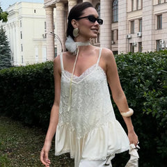 Woman in a white lace dress walking outdoors with greenery and a building in the background