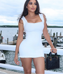 A woman wearing a black mini dress with white floral appliques on the shoulder straps, holding a yellow bag.


