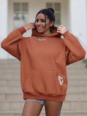 Woman wearing a brown hoodie with branding, standing outdoors.
