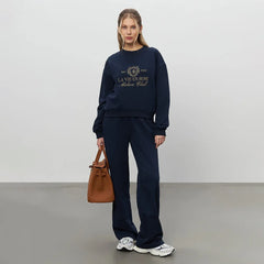 Woman wearing a navy blue sweatshirt and pants with a brown handbag on a plain background