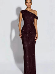A woman modeling a floor-length, one-shoulder evening gown covered in sparkling sequins. The dress is a wine color and is photographed against a plain light gray studio background with a soft shadow cast to the side.