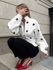 Person wearing a white blouse with black polka dots and black pants, squatting on steps.