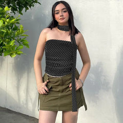Woman wearing a black polka dot top and green skirt with a white wall and plant in the background