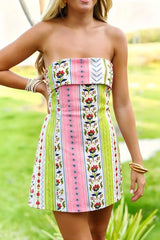 A woman wearing a strapless mini dress with vertical stripes in pink, lime green, white, and floral embroidery, standing outdoors on a grassy lawn.