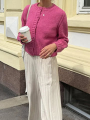 Person wearing a pink knitted cardigan and white pleated pants, holding a coffee cup outdoors.