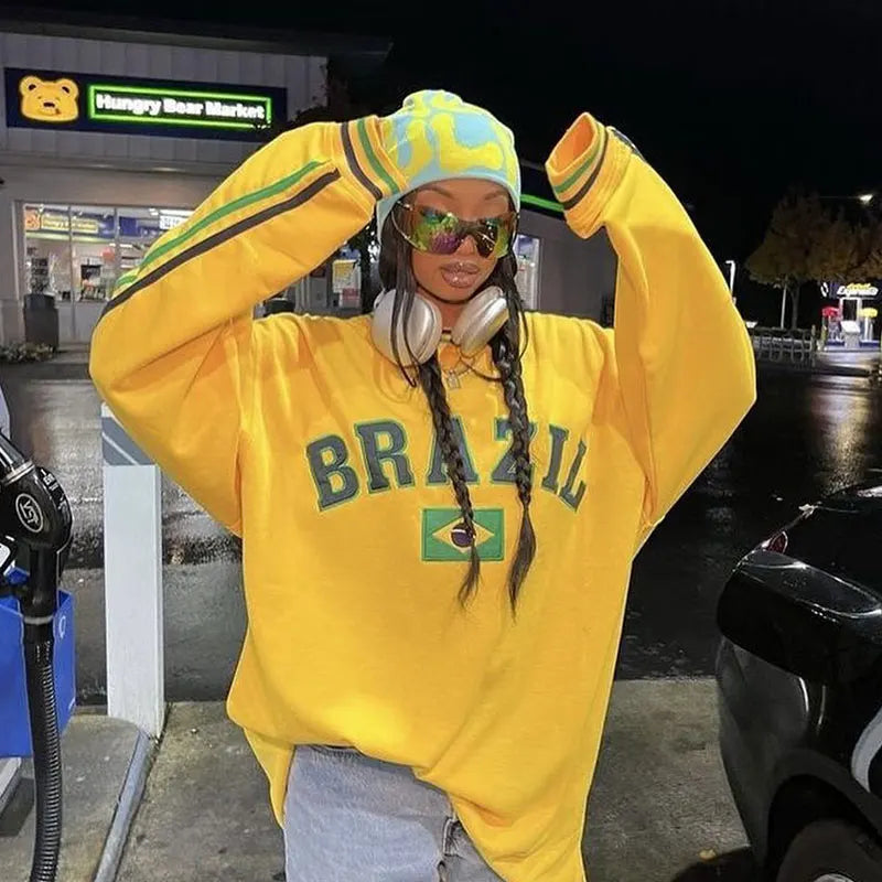 Person wearing a yellow 'Brazil' sweatshirt with braided hair, standing at a gas station.