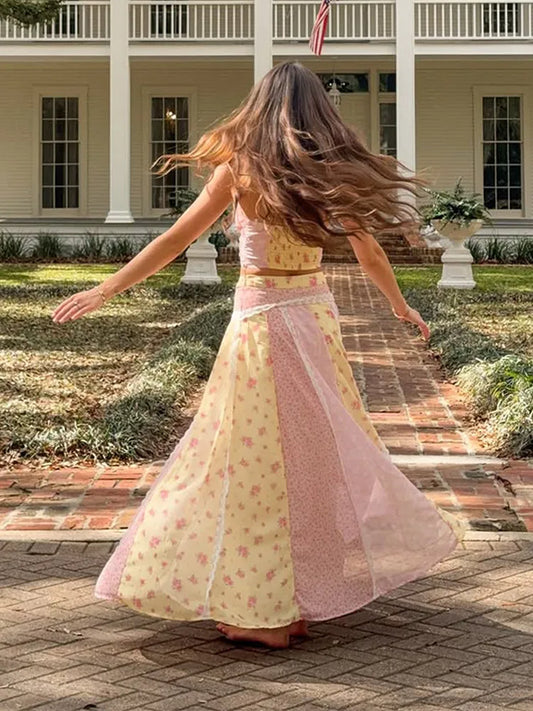 A woman with long brown hair is seen from behind, twirling in a pastel pink and yellow floral two-piece outfit with sheer panels, set against a backdrop of a white colonial-style house and brick pathway.