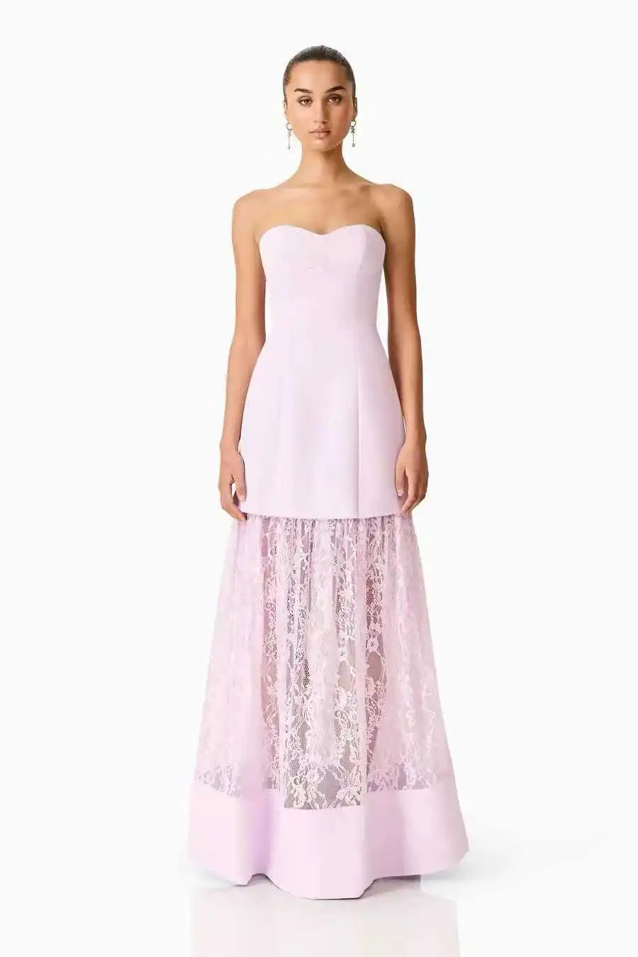 A woman modeling a strapless light pink gown with a structured sweetheart bodice and a full skirt featuring sheer floral lace overlay, photographed against a plain white studio background.