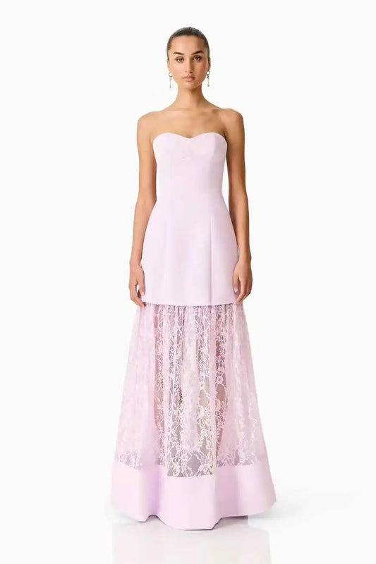 A woman modeling a strapless light pink gown with a structured sweetheart bodice and a full skirt featuring sheer floral lace overlay, photographed against a plain white studio background.