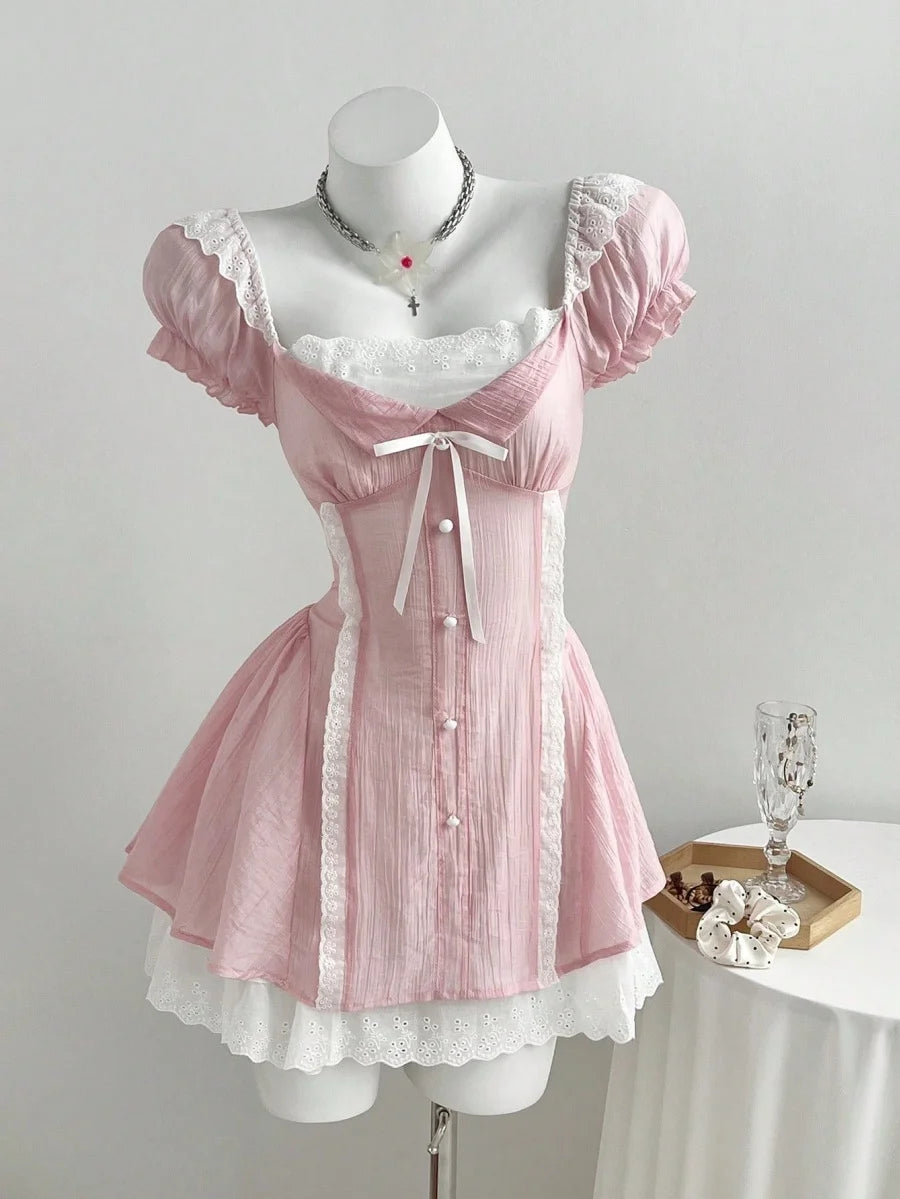 A light pink Lolita-style mini dress with white lace trim, puff sleeves, and a corseted bodice, displayed on a white mannequin. A small wooden tray with a glass and flowers sits on a white-covered table to the right, against a plain light gray wall.