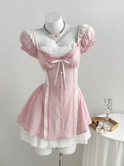 A light pink Lolita-style mini dress with white lace trim, puff sleeves, and a corseted bodice, displayed on a white mannequin. A small wooden tray with a glass and flowers sits on a white-covered table to the right, against a plain light gray wall.