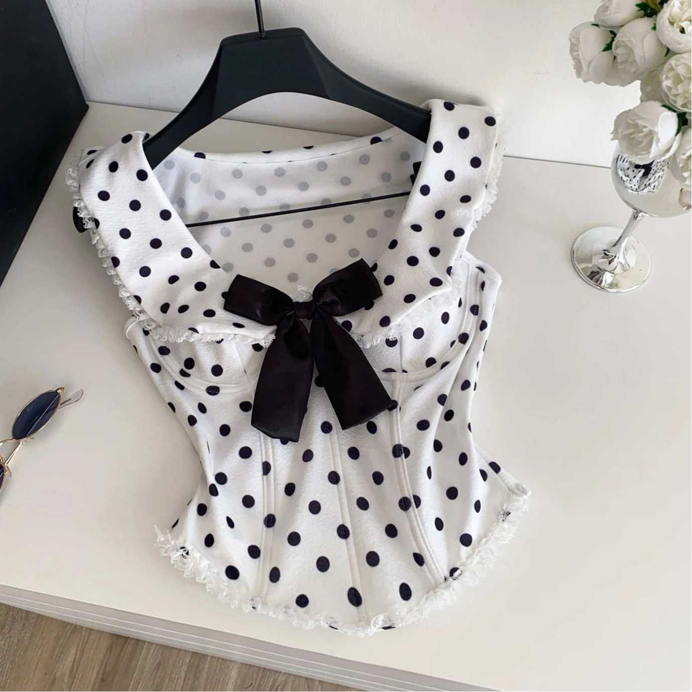 White blouse with black polka dots and a black bow on a white surface, with a  sunglasses in the background.