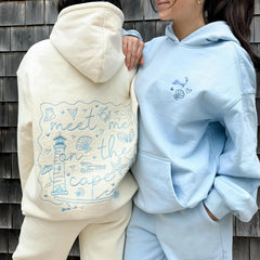 Two people wearing embroidered hoodies against a wooden wall.