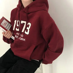 Person wearing a maroon hoodie with '1973' and 'New York' text, holding a book.