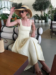 Woman in a yellow dress and sun hat sitting on a modern chair in an indoor setting with plants.