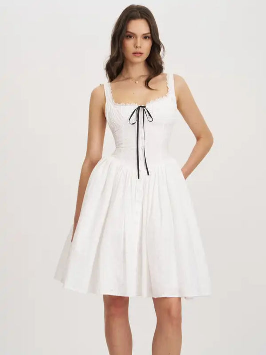 A woman modeling a white sleeveless fit-and-flare dress with a square neckline, delicate lace trim, a black center tie, and a flared skirt against a plain light background.