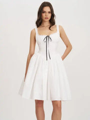 A woman modeling a white sleeveless fit-and-flare dress with a square neckline, delicate lace trim, a black center tie, and a flared skirt against a plain light background.