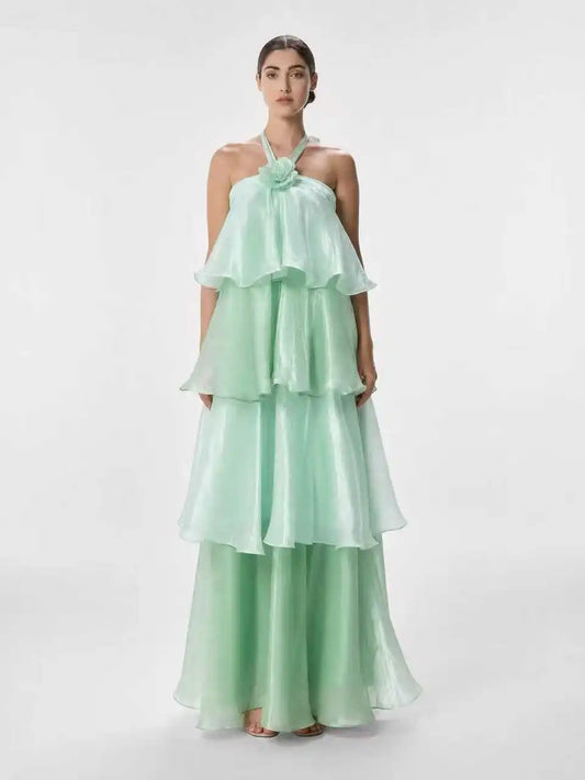 A woman modeling a mint green halter-neck maxi dress with a layered, tiered ruffle skirt and matching wide-leg pants against a plain light gray studio background.