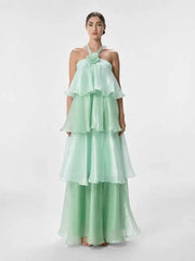 A woman modeling a mint green halter-neck maxi dress with a layered, tiered ruffle skirt and matching wide-leg pants against a plain light gray studio background.
