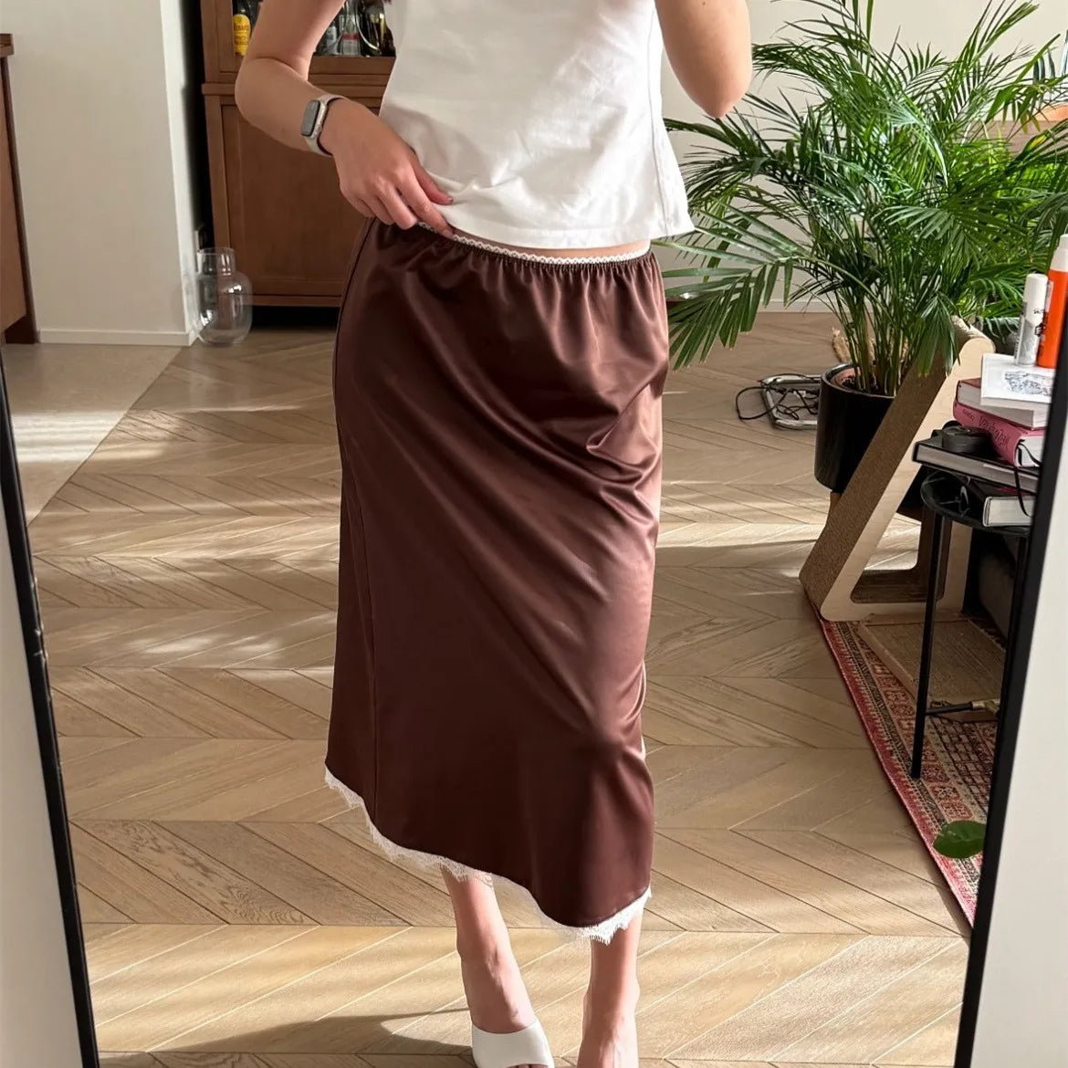 Person wearing a white top and brown skirt standing in a room with wooden flooring and plants.