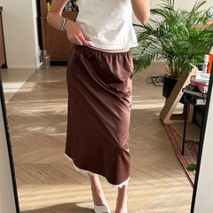 Person wearing a white top and brown skirt standing in a room with wooden flooring and plants.
