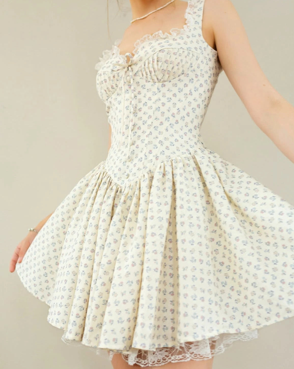 A sleeveless cream-colored swing dress with a small blue floral pattern, featuring a ruffled sweetheart neckline, lace trim, and a flared skirt, shown on a model against a neutral beige background.