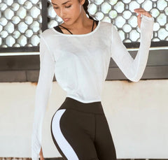 Woman wearing a white long-sleeve top and black leggings, standing against a textured wall.