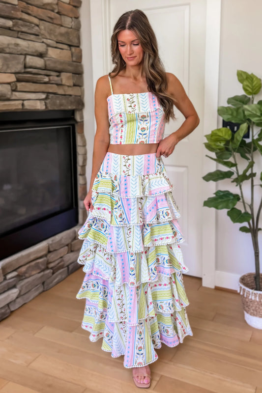 Woman wearing a colorful tiered dress standing in a room with a stone fireplace and a plant.